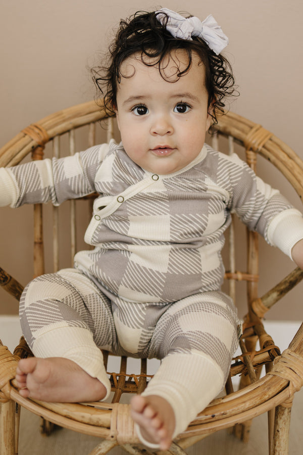 Baby in a checkered outfit sitting on a wicker chair with a neutral background